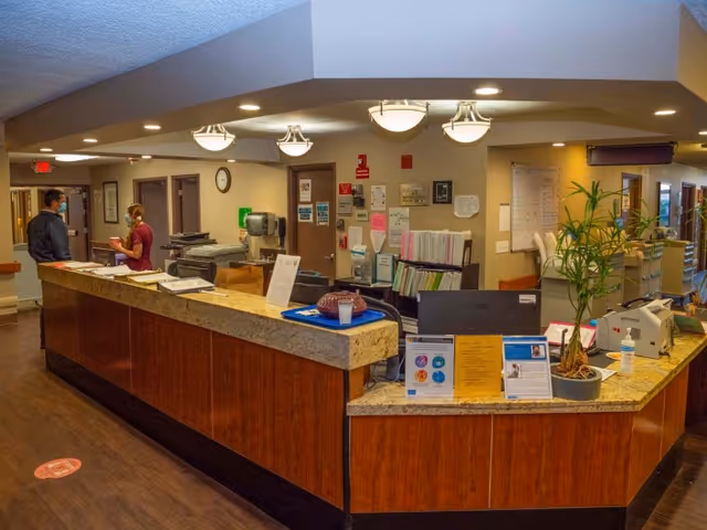 Reception area inside Somerset Subacute Rehab with a long wooden counter topped with granite. Two people are conversing near the counter. The area is well-lit with ceiling lights, and there are various office supplies, plants, and informational signs on the counter. The background shows doors, a clock, and wall-mounted documents.