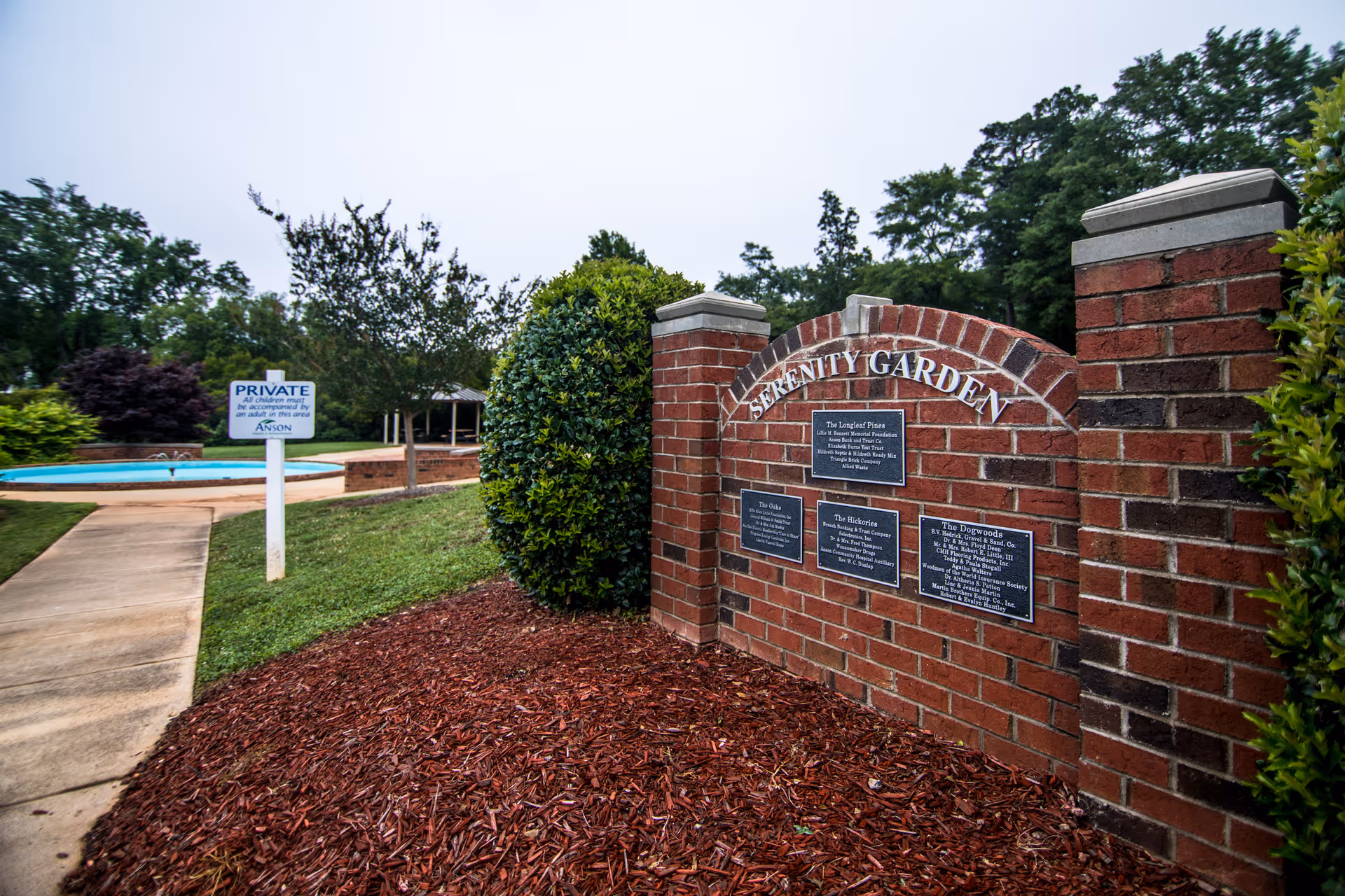 A landscaped outdoor area with a brick sign reading 'Serenity Garden' and plaques describing different types of plants. There is a paved walkway leading to a small pool or water feature, surrounded by trees and bushes under an overcast sky.