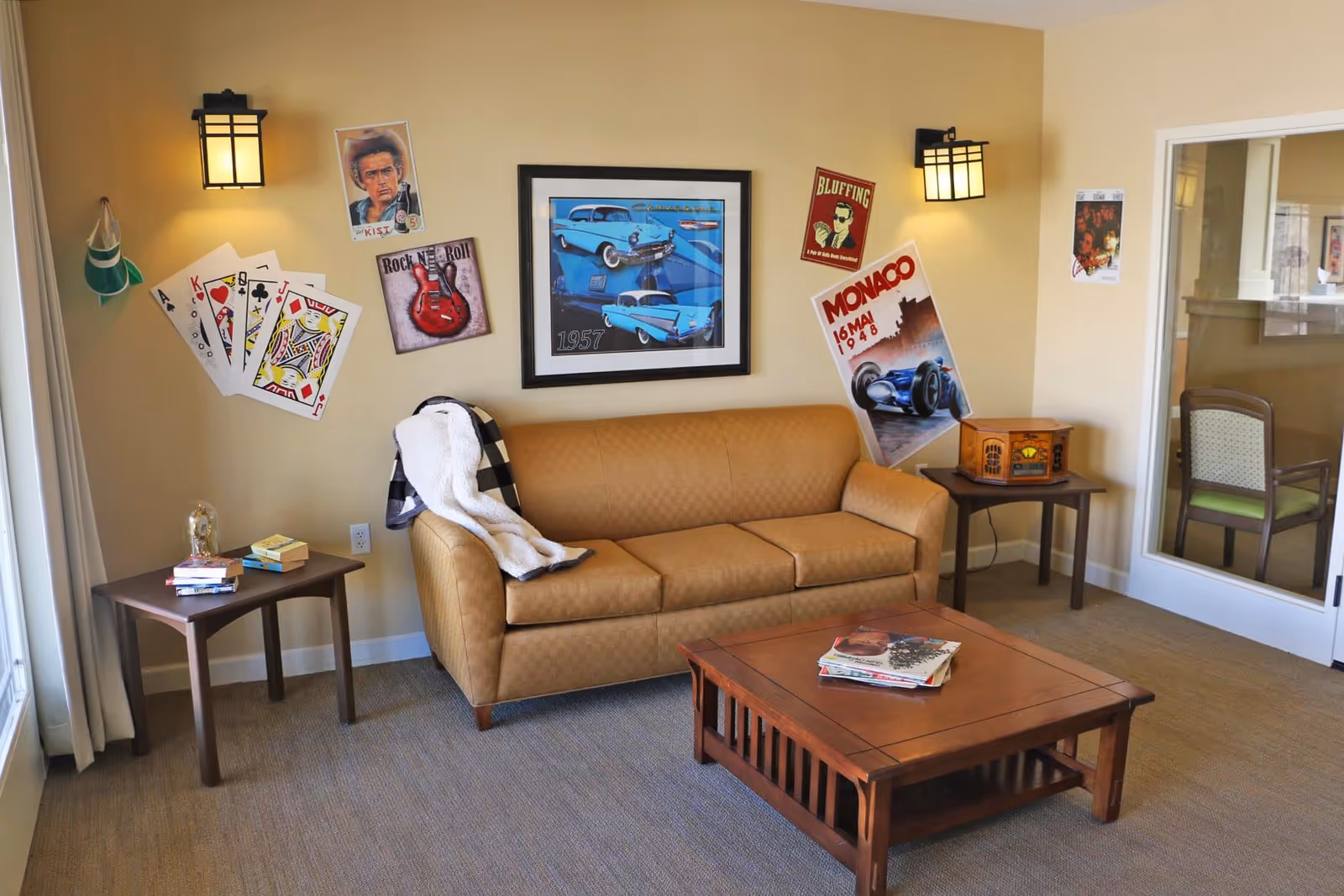 A cozy living room with a tan leather couch draped with a black and white blanket, a wooden coffee table with magazines, and two side tables. The walls are decorated with vintage posters, including playing cards, a guitar, a blue car from 1957, and a Monaco race car. Two wall-mounted lamps provide warm lighting, and a glass door reveals another room with chairs.