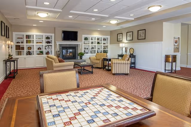 A spacious common area with a patterned carpet, several upholstered chairs and sofas arranged around a coffee table and fireplace. Built-in white shelves with decorative items flank the fireplace, and a flat-screen TV is mounted above it. A Scrabble game board is visible on a wooden table in the foreground. The walls are light-colored with framed artwork and lamps providing soft lighting.
