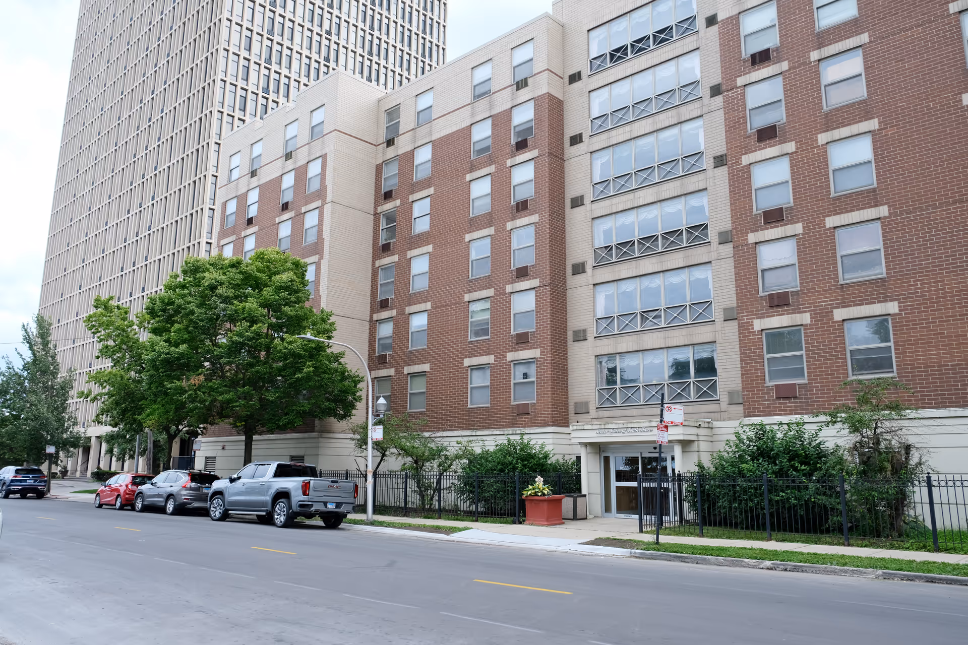Exterior view of a multi-story senior living facility building with a combination of beige and red brick facade, several windows, and an entrance door. There are trees, parked cars along the street, and a sidewalk in front of the building.