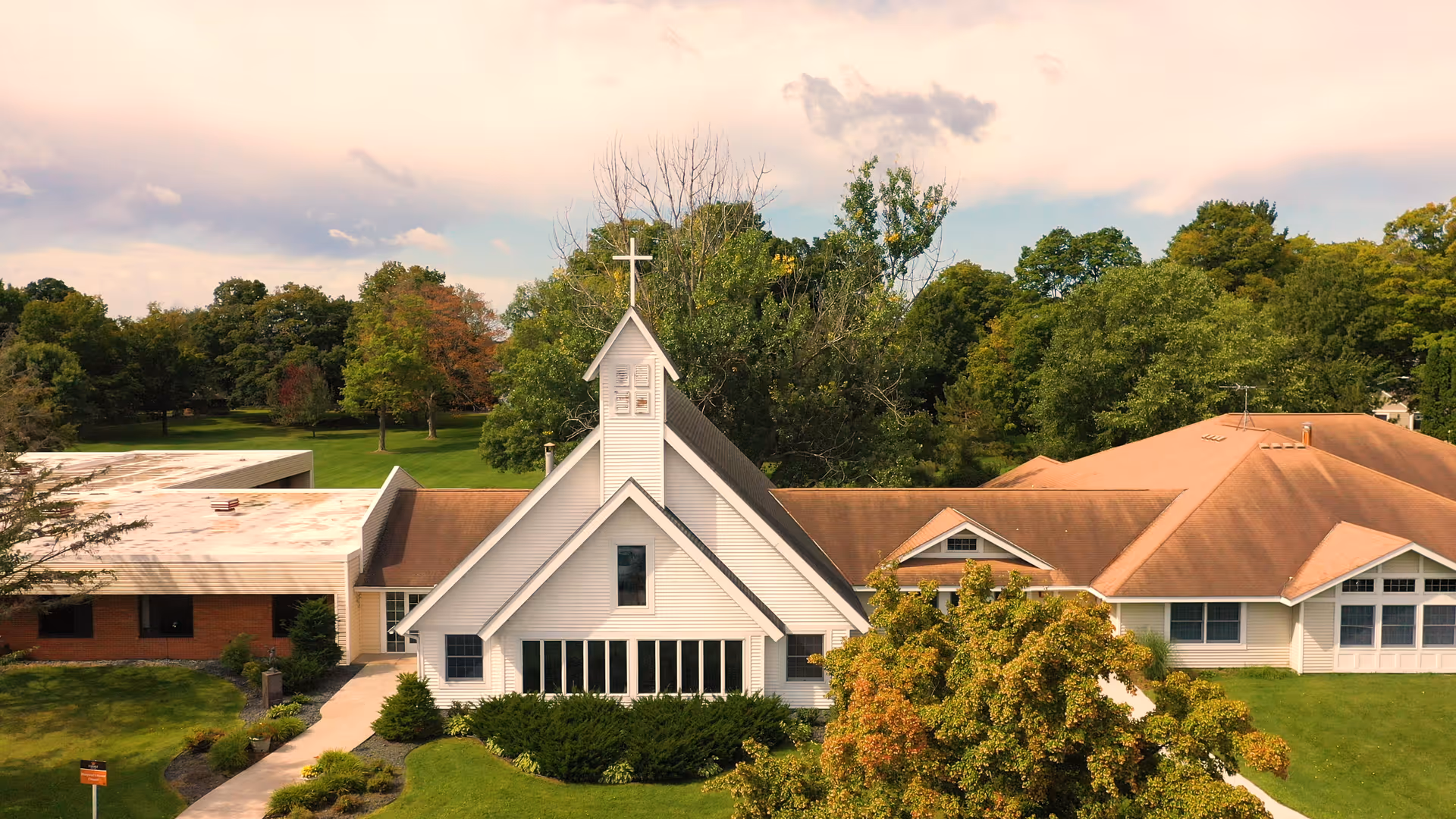 Front exterior view of a white chapel-style building with a cross on the steeple, flanked by attached buildings and surrounded by lawns and trees.