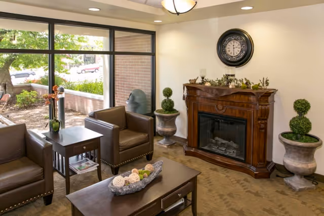 A cozy living room area with two brown leather armchairs and a matching sofa around a dark wooden coffee table decorated with a centerpiece of decorative balls. There is a wooden fireplace with a clock above it on the wall, flanked by two large potted plants. Large windows show an outdoor garden area with greenery and a brick wall.