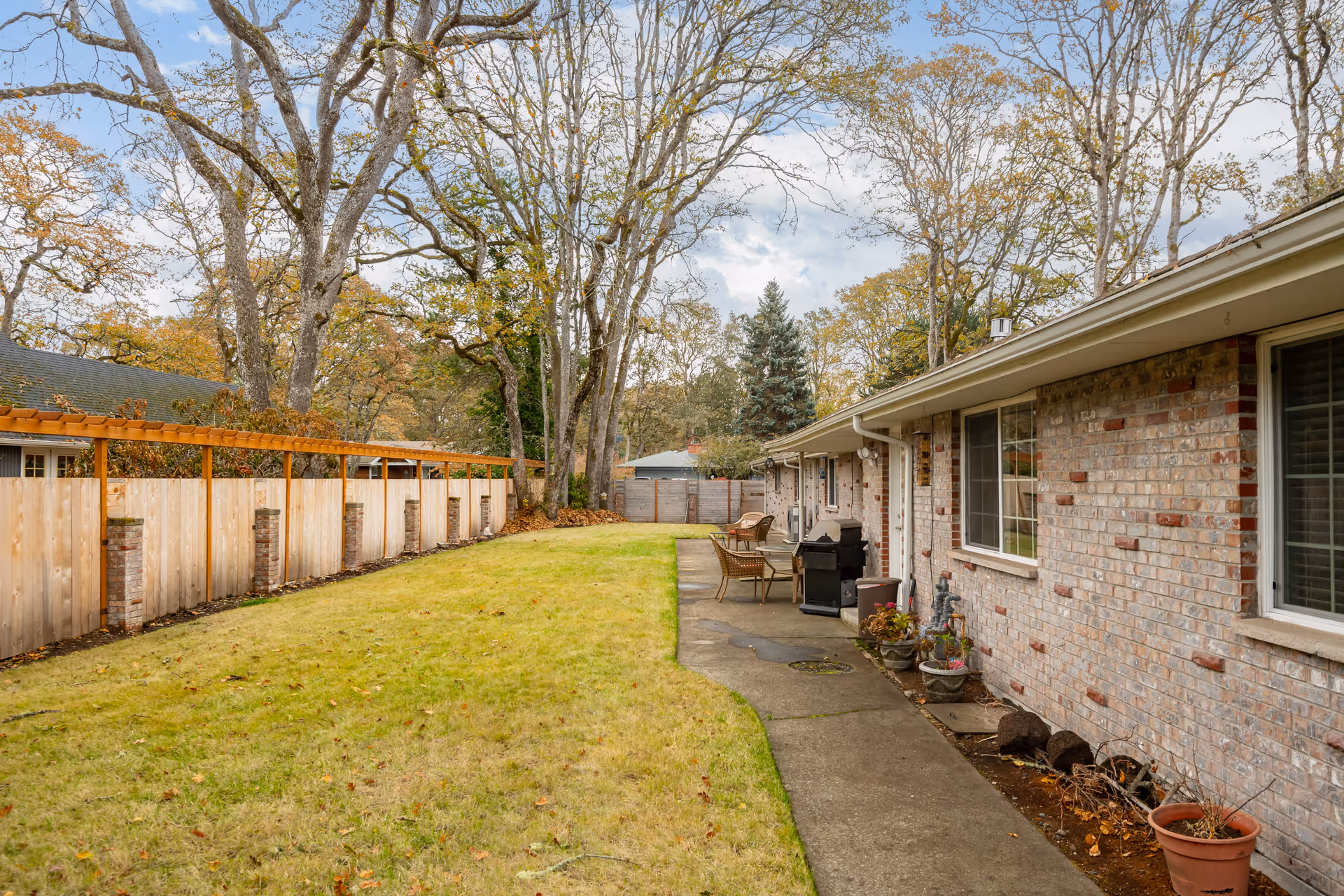Rear exterior of a single-story brick home with a lawn, concrete walkway, patio furniture, and a wooden fence backed by tall trees.