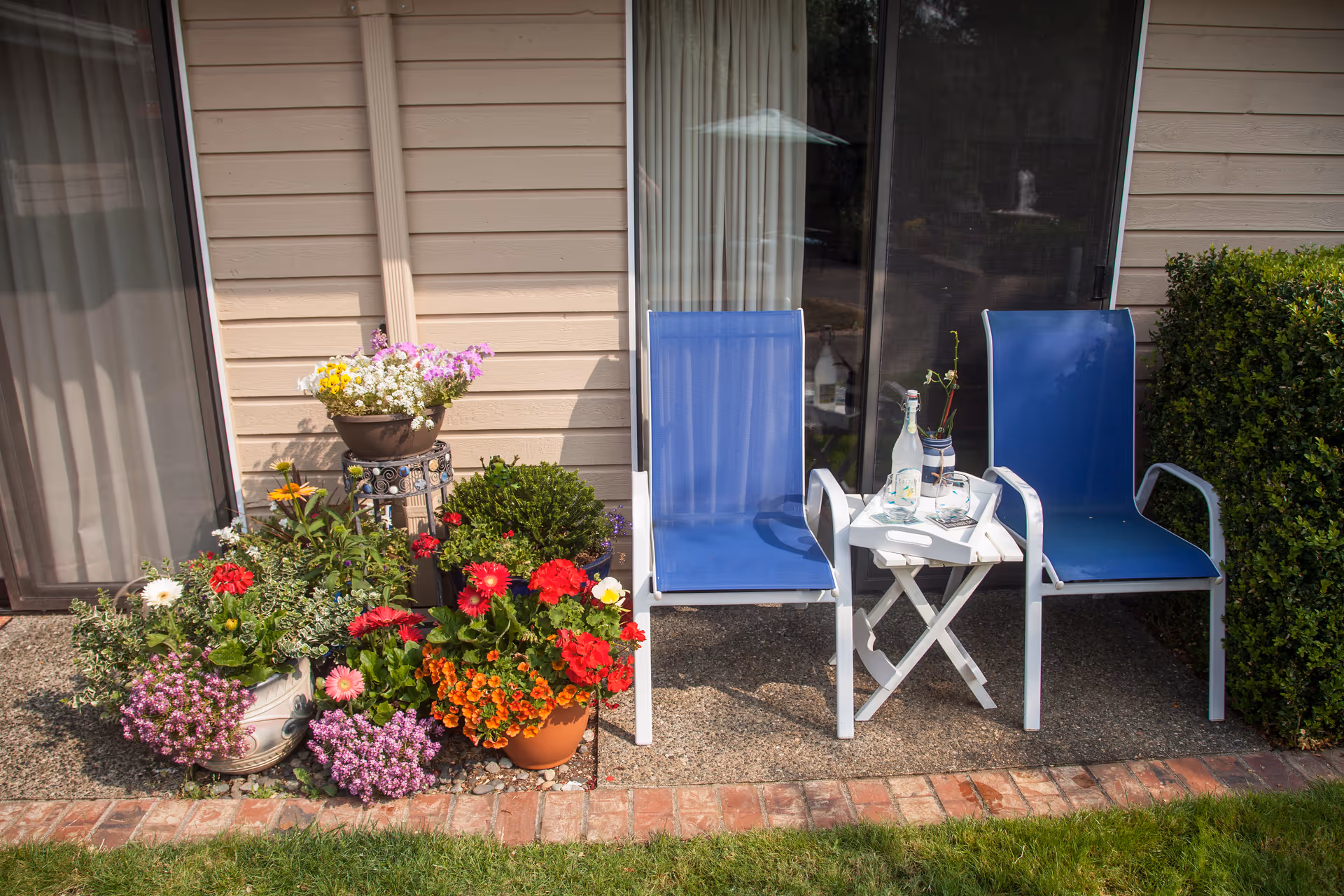 Two blue patio chairs with white frames sit on a concrete patio next to a small white folding table holding a bottle, a glass, and a small plant. To the left of the chairs is a collection of colorful potted flowers and greenery. The patio is adjacent to a beige siding building with sliding glass doors and a window covered by curtains. A green hedge is visible on the right side.