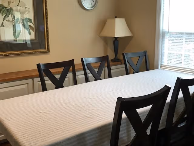 Dining room with a rectangular table covered by a white tablecloth surrounded by six dark wooden chairs. A table lamp with a beige shade sits on a sideboard against the wall, which also features a framed floral painting and a round wall clock. A window with blinds allows natural light into the room.