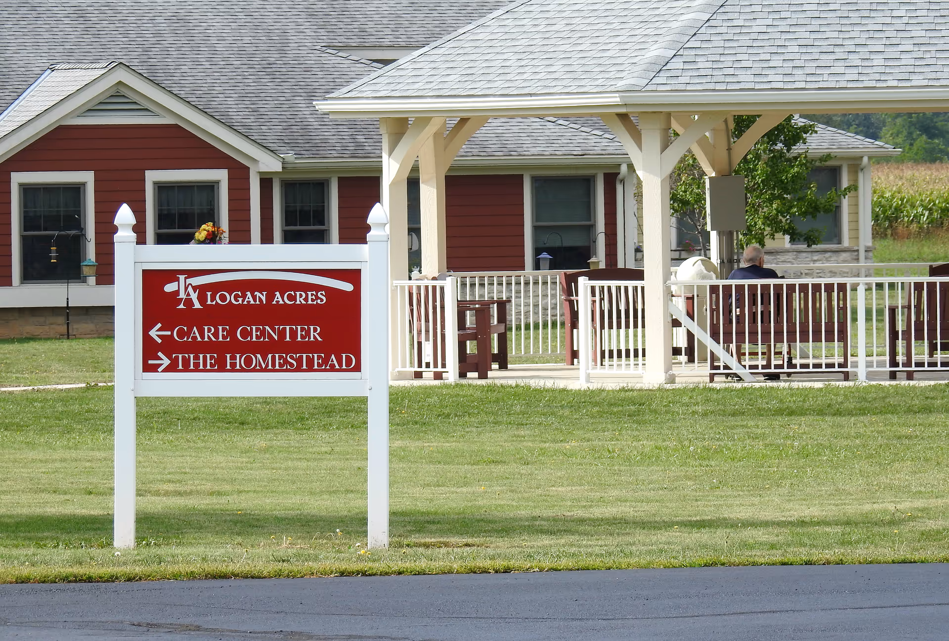 A white sign reading "Logan Acres" stands on a lawn in front of a red building and a covered outdoor seating area.