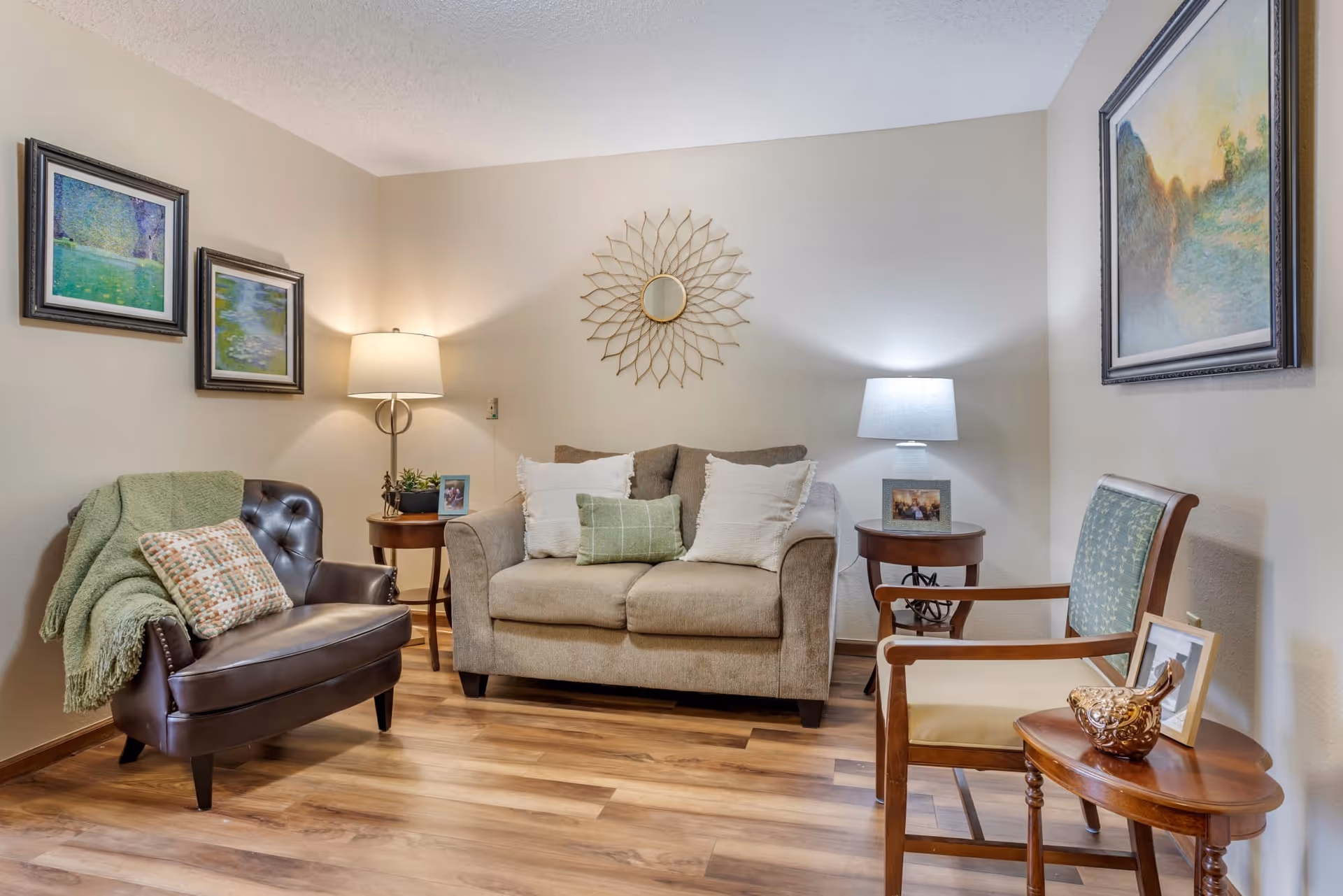 A cozy living room with a beige loveseat adorned with three pillows, a dark brown leather armchair with a green throw blanket and patterned pillow, and a wooden armchair with a green cushion. There are two round wooden side tables with lamps and framed photos. The walls are decorated with framed paintings and a decorative sunburst mirror. The floor is wood laminate.