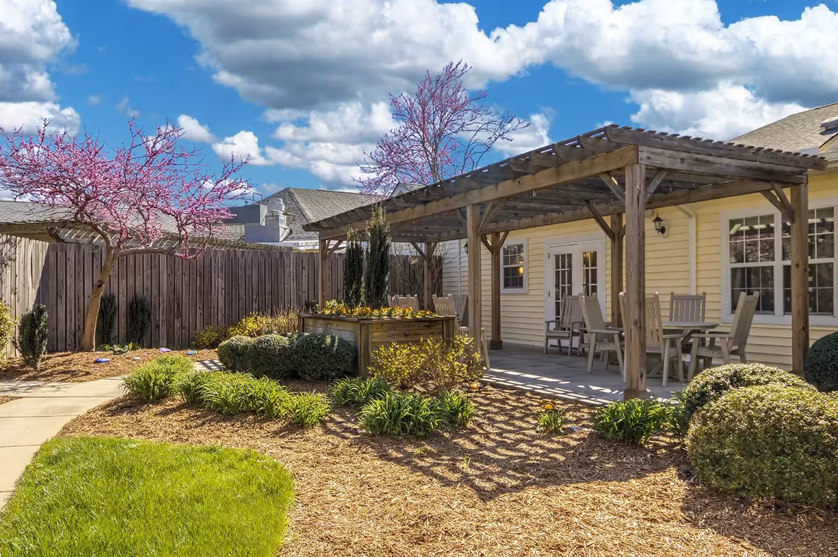 Outdoor patio area at TerraBella Shelby featuring a wooden pergola with tables and chairs underneath, surrounded by landscaped garden beds with green shrubs and flowering trees, under a partly cloudy blue sky.