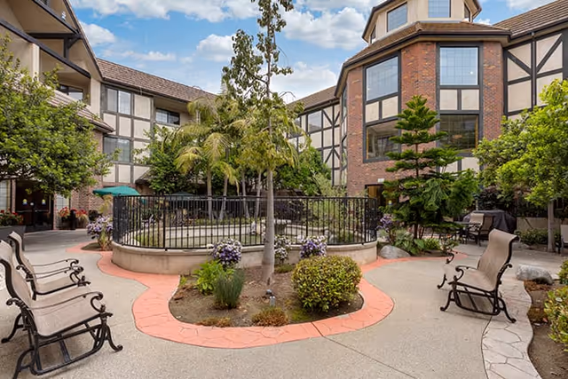 Outdoor courtyard area at Brookdale Brookhurst featuring a circular fenced garden with plants and trees, surrounded by benches and chairs. The courtyard is enclosed by a multi-story building with large windows and Tudor-style architecture under a partly cloudy sky.