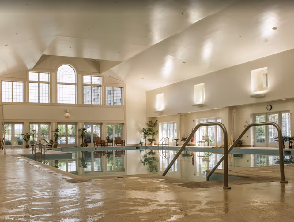 A spacious indoor pool area at Garden Spot Village, featuring large windows that allow natural light, surrounded by plants and seating.