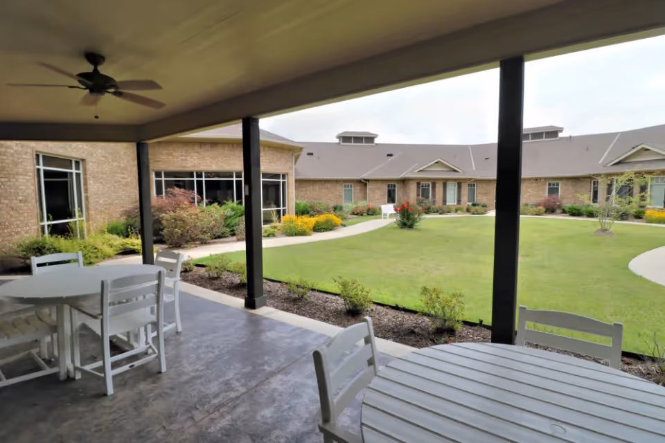 Covered patio with white tables and chairs overlooking a grassy courtyard and brick senior living building.