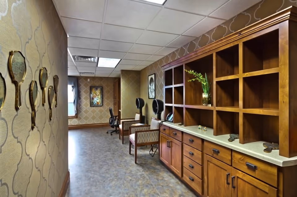 A hallway in a senior living facility with patterned wallpaper and a row of decorative hand mirrors mounted on the left wall. On the right side, there is a large wooden cabinet with multiple open cubbies and drawers, decorated with a vase containing green plants. Further down the hallway, there are chairs and two black hair dryer chairs against the wall, with framed artwork hanging above them.