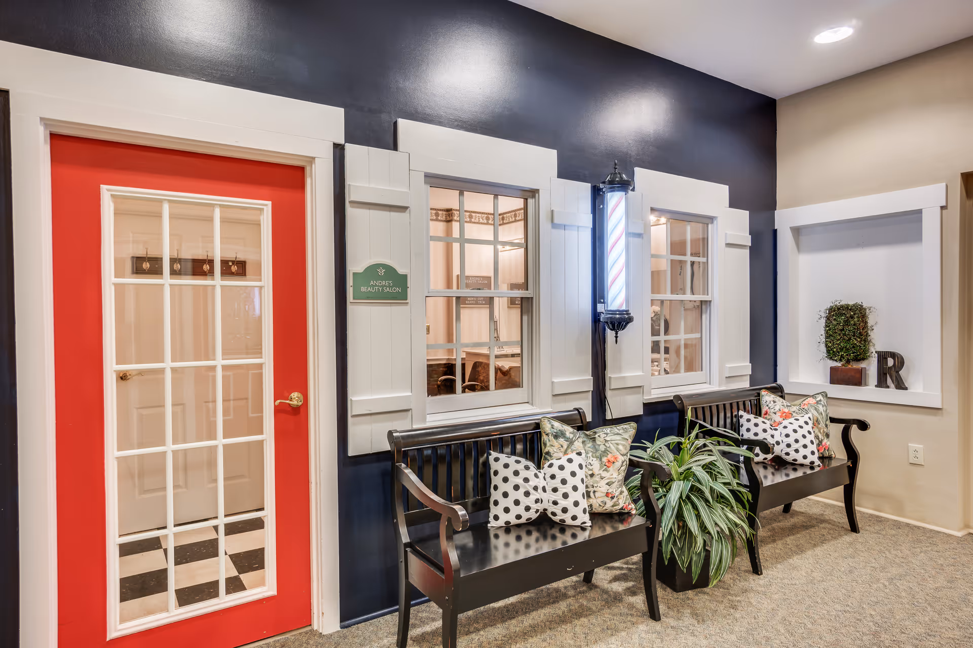 Decorative interior seating area with two black benches, patterned pillows, a potted plant, faux windows, a red door and a barber pole.