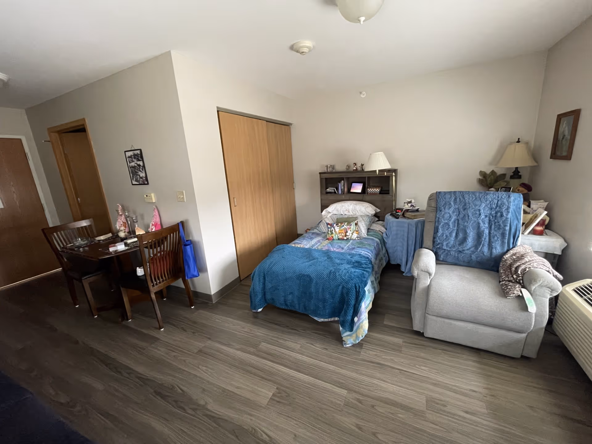 A small, cozy room with a single bed covered in a blue blanket and colorful pillows. Next to the bed is a gray recliner chair with a blue throw draped over the back. A small table with a lamp and various items is positioned between the bed and chair. On the left side of the room, there is a wooden dining table with two chairs and some decorative items on top. The room has wood flooring and light-colored walls, with a closet featuring sliding wooden doors.