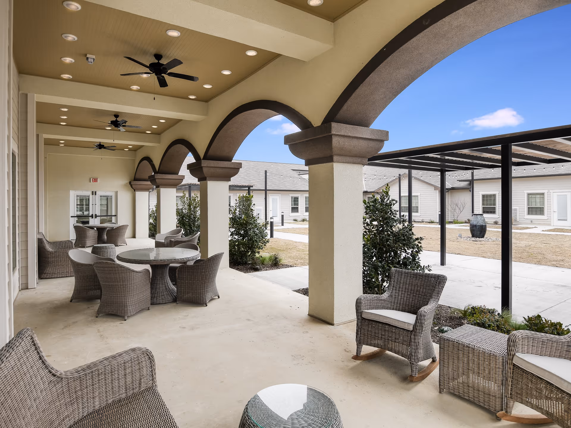 Covered outdoor patio area with wicker chairs, round tables, ceiling fans, and arched columns overlooking a courtyard with shrubs and a water feature under a clear blue sky.