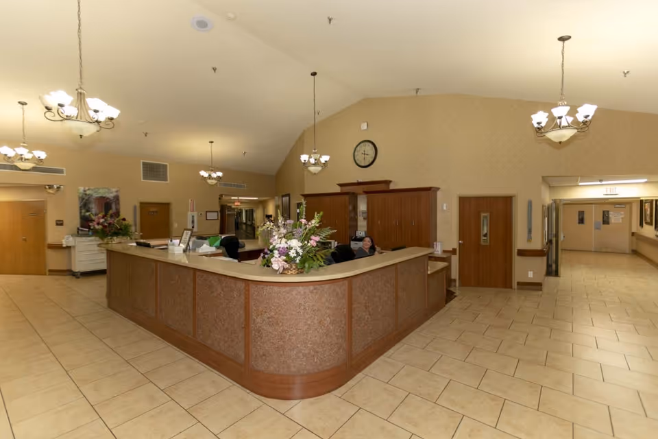 Reception area inside a senior living facility with a large curved wooden front desk adorned with flower arrangements. Behind the desk, a staff member is seated. The room has beige tiled floors, beige walls, multiple wooden doors, and several ceiling light fixtures. A clock is mounted on the wall above the desk.