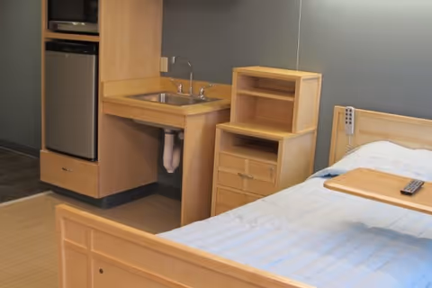 A senior living facility room featuring a wooden bed with a light blue bedspread, a wooden bedside table with shelves and drawers, a small sink with a faucet, and a mini refrigerator built into wooden cabinetry.