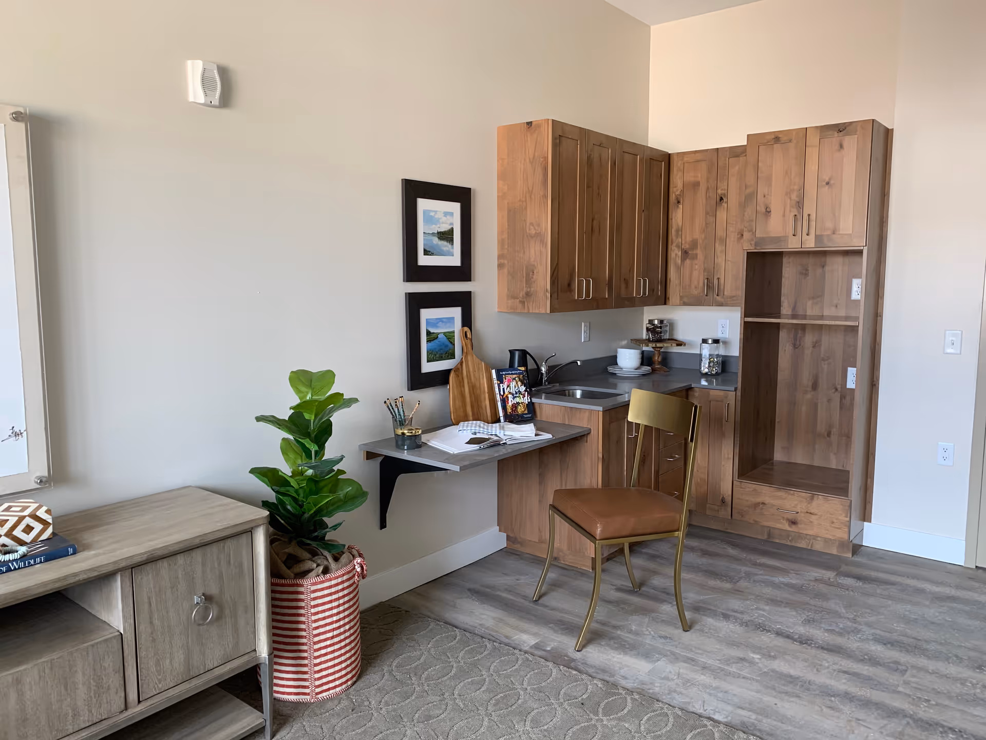A small kitchenette area with wooden cabinets, a sink, and a countertop. A single chair with a brown seat and gold frame is placed in front of a fold-down wall-mounted table with a notebook, pencils, and a book on it. Two framed landscape pictures hang on the wall above the table. To the left, there is a green potted plant in a red and white striped basket next to a wooden cabinet with a decorative pillow and a book on top. The floor has a wood-like finish and a patterned rug.