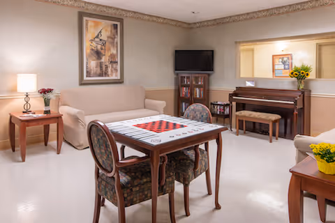 A cozy common area in a senior living facility featuring a beige couch, a wooden table with a checkerboard game, four patterned chairs, a piano with a bench, a small bookshelf with a TV on top, and side tables with lamps and flower arrangements. The walls are decorated with framed artwork and the floor is light-colored.