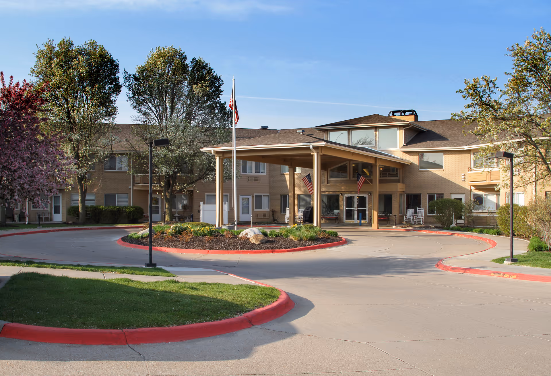 Exterior view of Asher Point Independent Living of Lincoln showing the front entrance with a covered driveway, landscaped roundabout with plants and rocks, several trees, and an American flag on a flagpole under a clear blue sky.