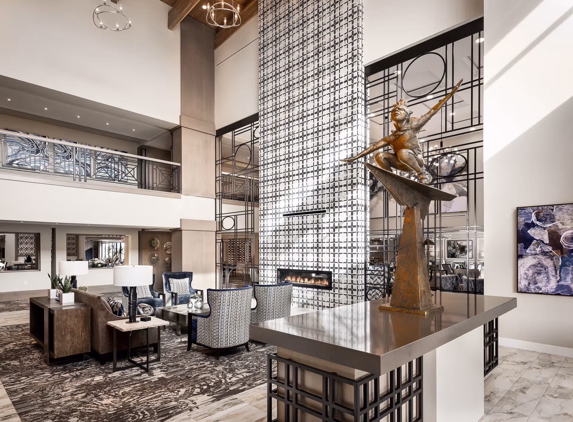 A spacious and modern living room area with high ceilings, featuring a patterned tile fireplace, comfortable seating including armchairs and a sofa, decorative lamps, and a bronze sculpture of a child on a paper airplane on a polished table. The room has a neutral color palette with accents of blue and intricate metalwork railings on the upper level.