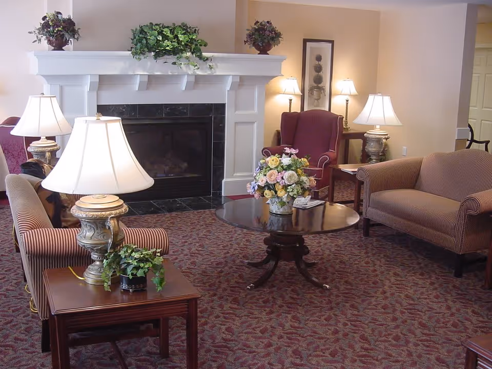 A cozy living room area with a fireplace in the center, flanked by two table lamps on side tables. The room features a patterned carpet, a round wooden coffee table with a floral arrangement, a striped loveseat, and a burgundy armchair. The walls are light-colored with framed artwork and additional lamps in the background.