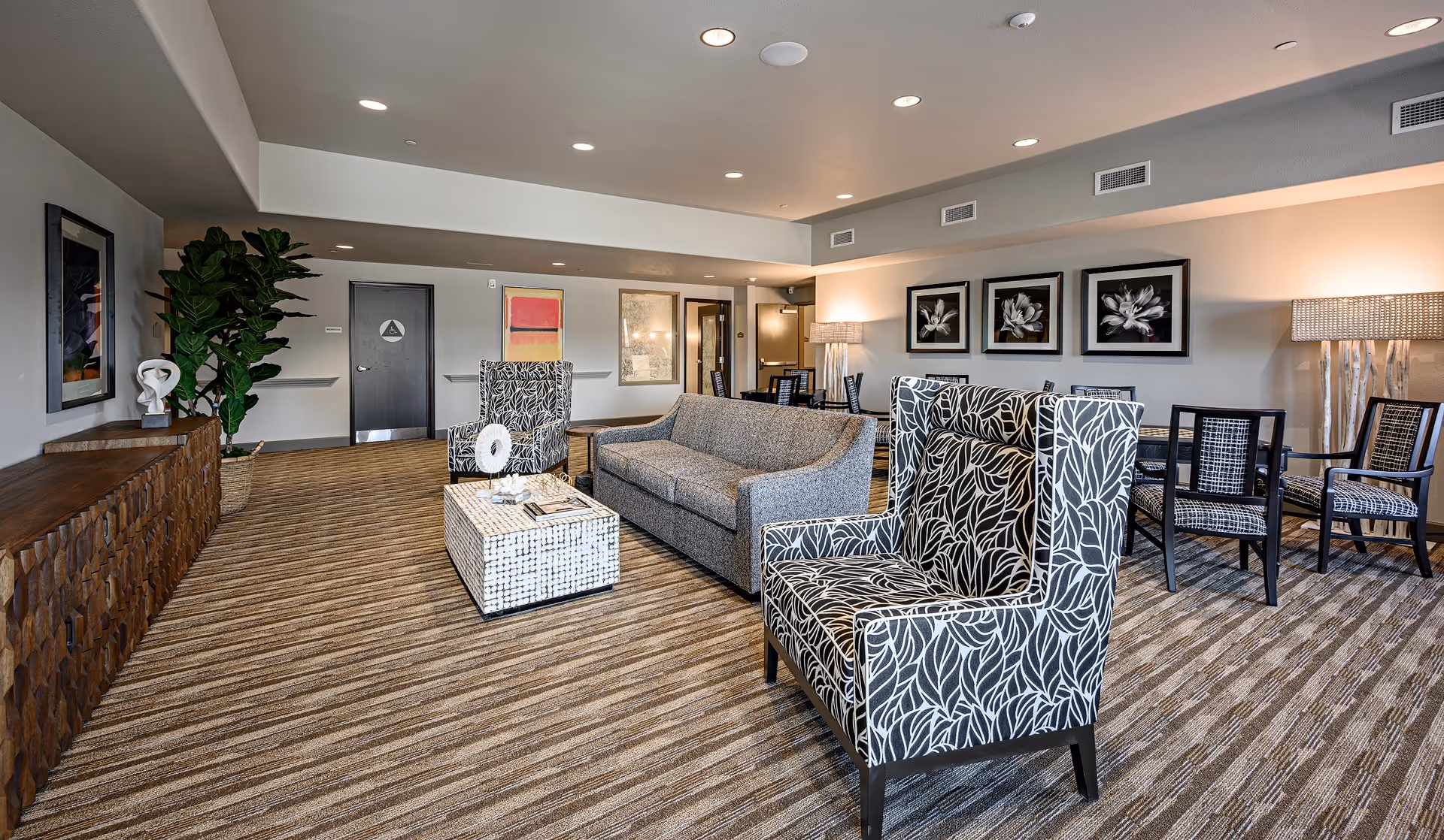 A spacious and well-lit living room area in a senior living facility featuring a gray sofa, two patterned armchairs with black and white leaf designs, a white textured coffee table with decorative items, a wooden sideboard with a sculpture and a large potted plant, and a dining area with multiple chairs and tables. The walls are adorned with framed artwork and the room has recessed ceiling lights and carpeted flooring.