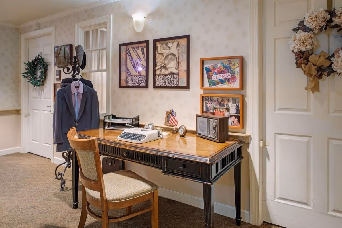 A vintage-style wooden desk with a typewriter, small American flag, clock, and radio on top. A wooden chair with a cushioned seat is placed in front of the desk. On the wall above the desk are framed pictures and artwork. To the left, a coat rack holds a suit jacket and hats. The room has beige wallpaper with a subtle pattern, carpeted floor, and two white doors decorated with wreaths.