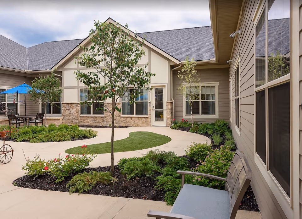 Outdoor courtyard area at StoryPoint Shaker Heights featuring a curved concrete walkway, small trees, green shrubs, and flowering plants. There is a bench with a blue cushion on the right side and a table with chairs and a blue umbrella on the left side near the building. The building exterior has beige siding, stone accents, and multiple windows.