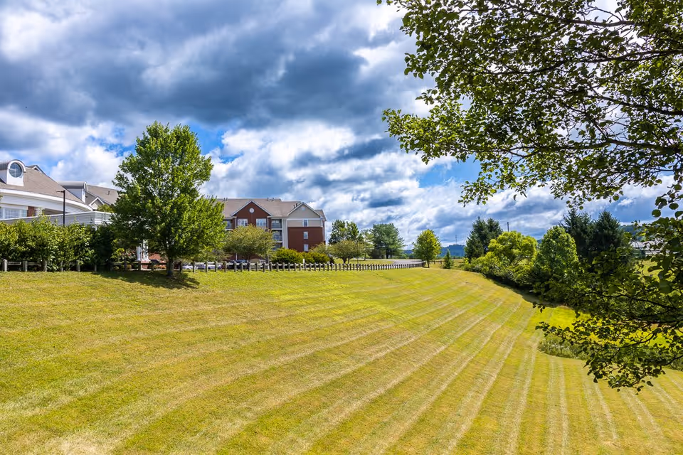 A large grassy area with neatly mowed stripes in front of a multi-story residential building partially visible on the left side. Trees and bushes border the grassy slope, and the sky above is partly cloudy with patches of blue.