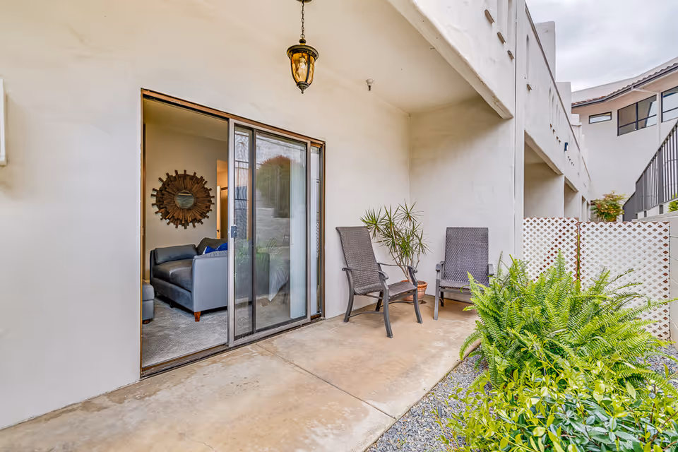 Outdoor patio area with two brown wicker chairs and potted plants, adjacent to a sliding glass door leading into a living room with a gray sofa and decorative wall mirror.