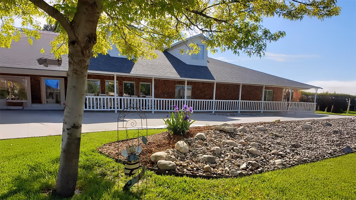 Exterior view of a single-story building with a gray roof and brick walls, featuring a white railing along a covered porch. In the foreground, there is a landscaped garden area with rocks, mulch, green grass, a tree, and a decorative bee ornament. The sky is clear and blue.