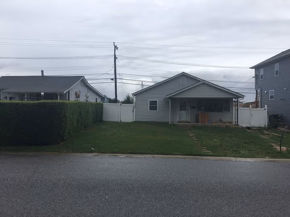 Front view of a single-story gray house with a small covered porch, a green lawn, and a white fence on a cloudy day.