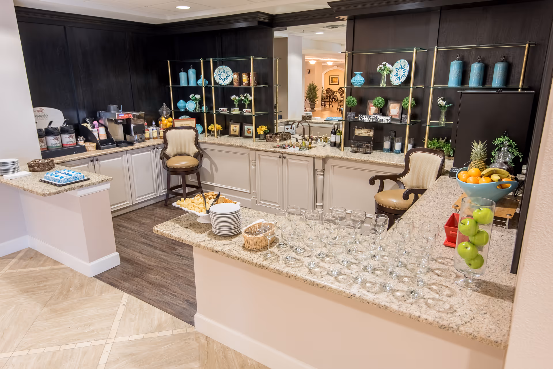 A well-lit kitchen area with granite countertops, two upholstered chairs, shelves displaying decorative plates, vases, and plants. The counter has a variety of glasses, plates, a bowl of fruit, and a tray with snacks. Coffee and beverage dispensers are visible on the left side.
