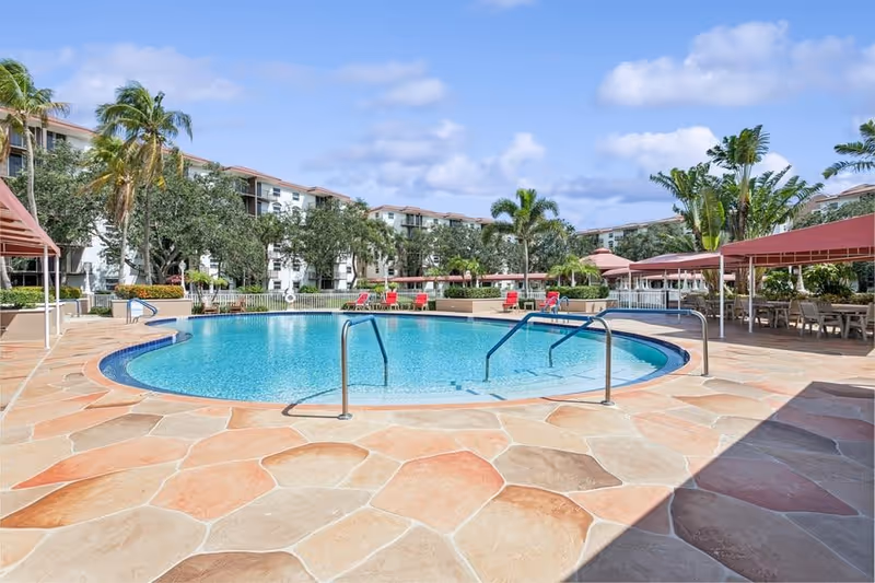 Outdoor swimming pool area with clear blue water surrounded by a stone tiled deck. There are metal handrails for pool entry, red lounge chairs, palm trees, and covered seating areas with tables and chairs. In the background, there are multi-story residential buildings and a partly cloudy sky.