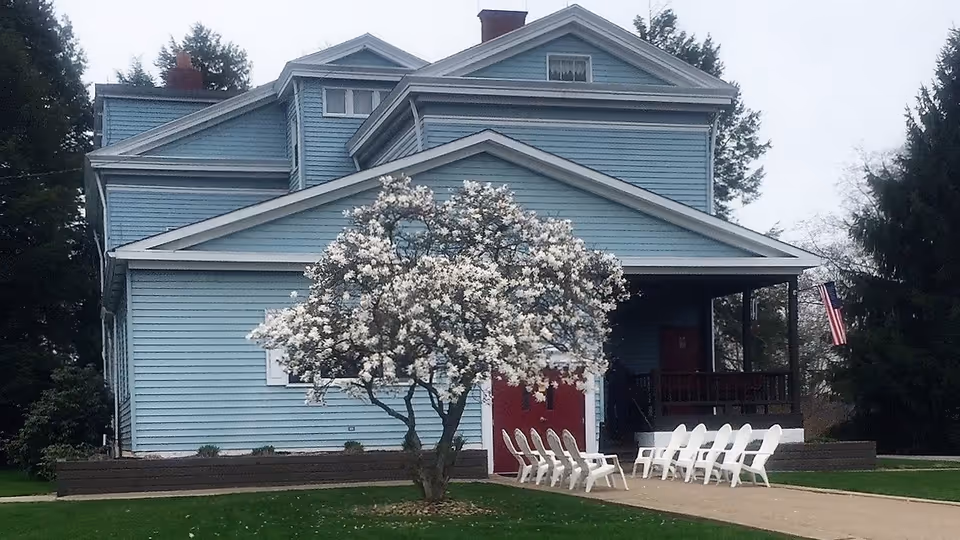 Exterior view of a large blue multi-story house with white trim and a red door. In front of the house is a blooming tree with white flowers and a row of white chairs arranged on a paved area. There are tall trees surrounding the property and an American flag is visible near the porch.