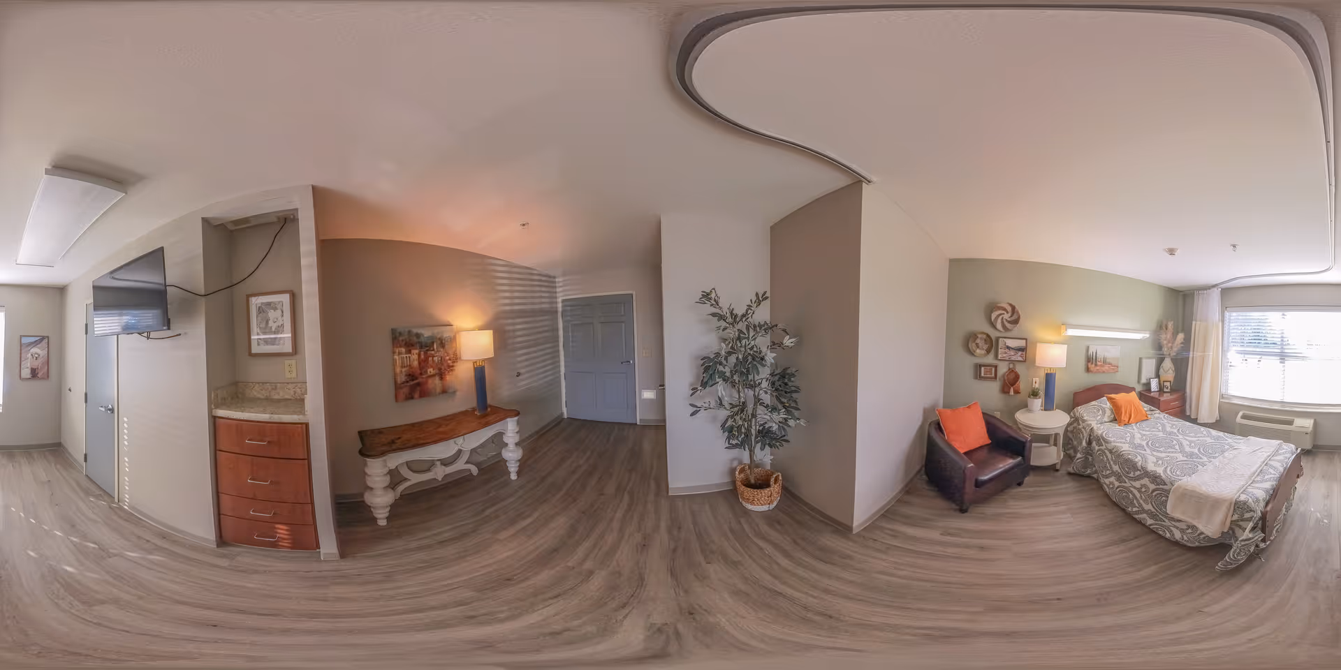 Bright assisted-living bedroom suite with a bed, armchair, bedside tables and lamp, TV on a dresser, and an entry hallway with a decorative table and potted plant.
