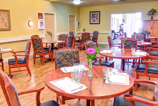 Bright communal dining room with round wooden tables set with placemats, glasses, and floral centerpieces.