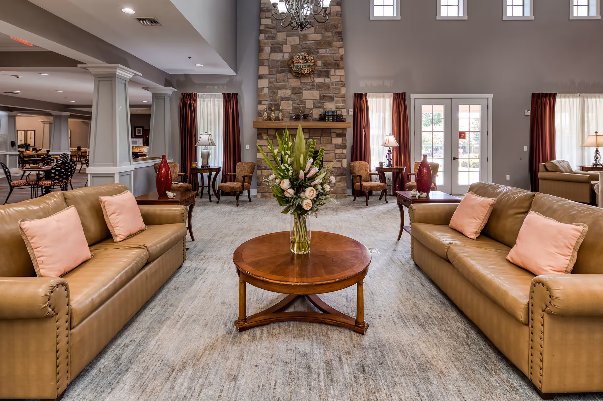 A spacious and well-lit living room area with two tan leather sofas facing each other, each adorned with pink cushions. A round wooden coffee table with a floral arrangement sits between the sofas. The room features a stone fireplace with a 'Welcome' sign above it, flanked by windows with red curtains and side tables with lamps. In the background, there are additional seating areas and a glass door leading outside.