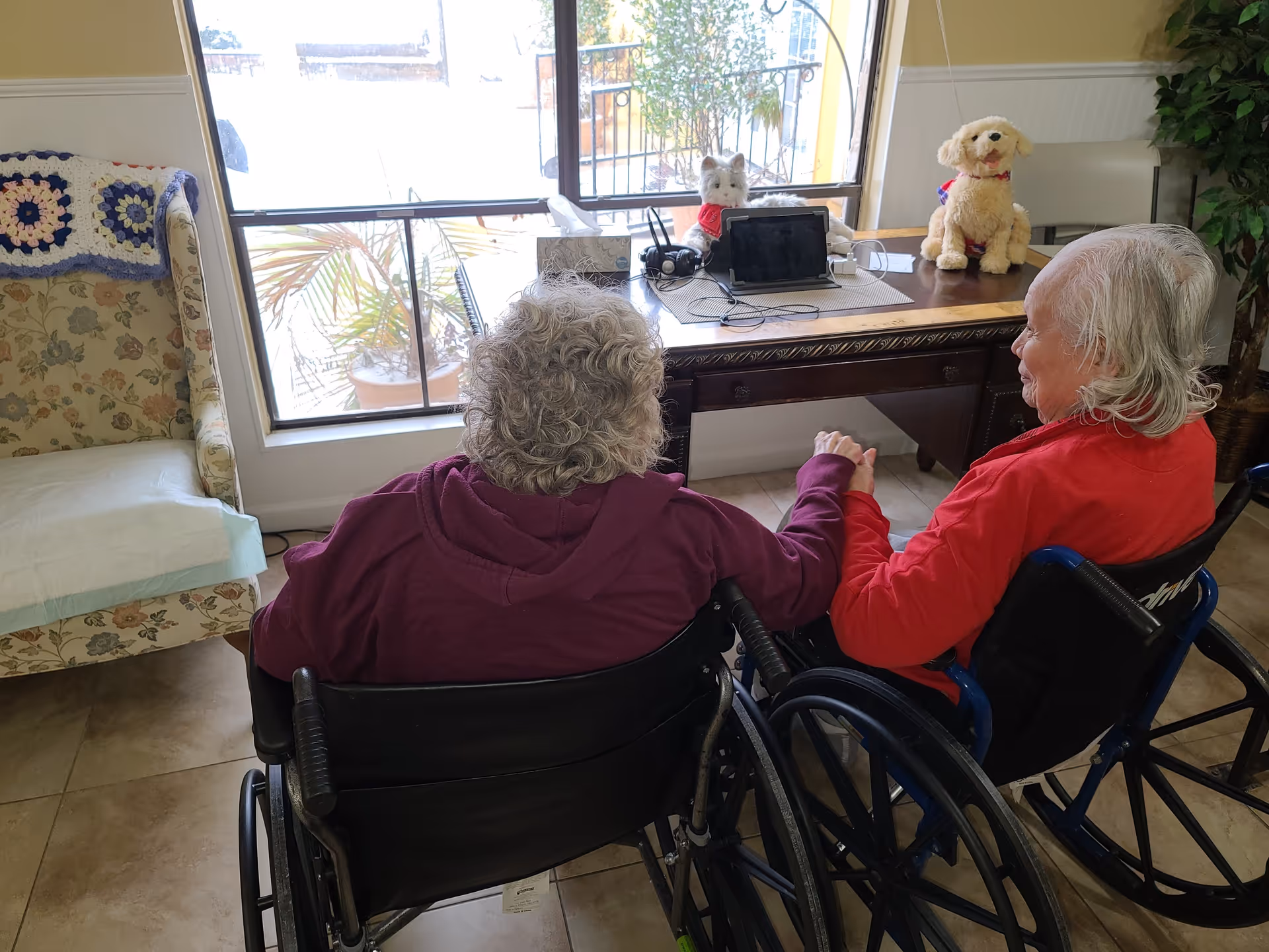 Two elderly individuals sitting in wheelchairs holding hands in a room with a window. There is a wooden table in front of them with a tablet, headphones, a tissue box, and two stuffed animals. A floral armchair with a crocheted blanket is to the left, and a potted plant is visible on the right.