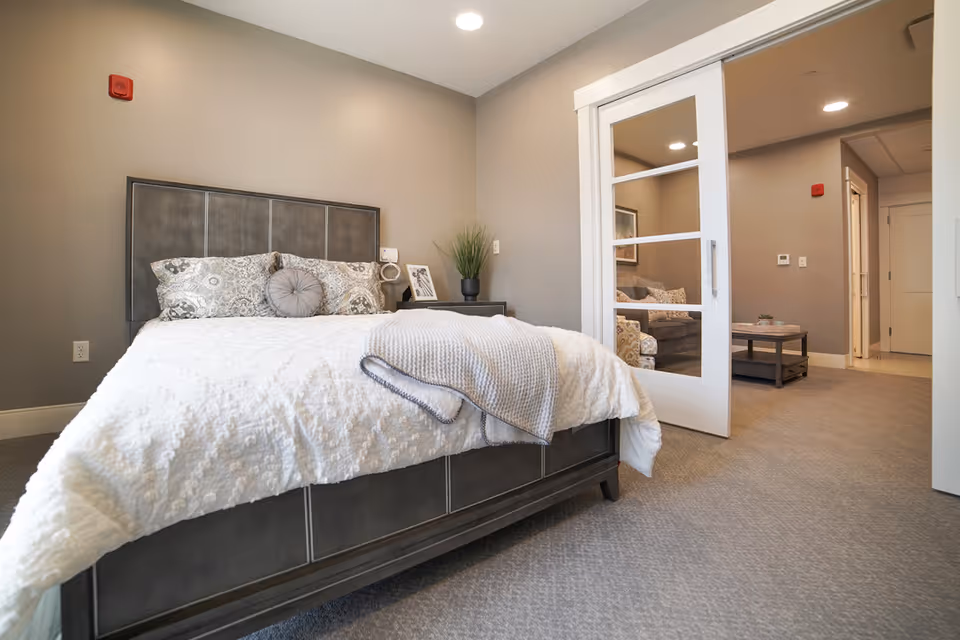A neatly made bed with patterned pillows and a gray throw blanket in a bedroom with beige walls and carpeted floor. A nightstand with a plant and framed photo is beside the bed. A sliding door with glass panels opens to a living area with a sofa and coffee table.