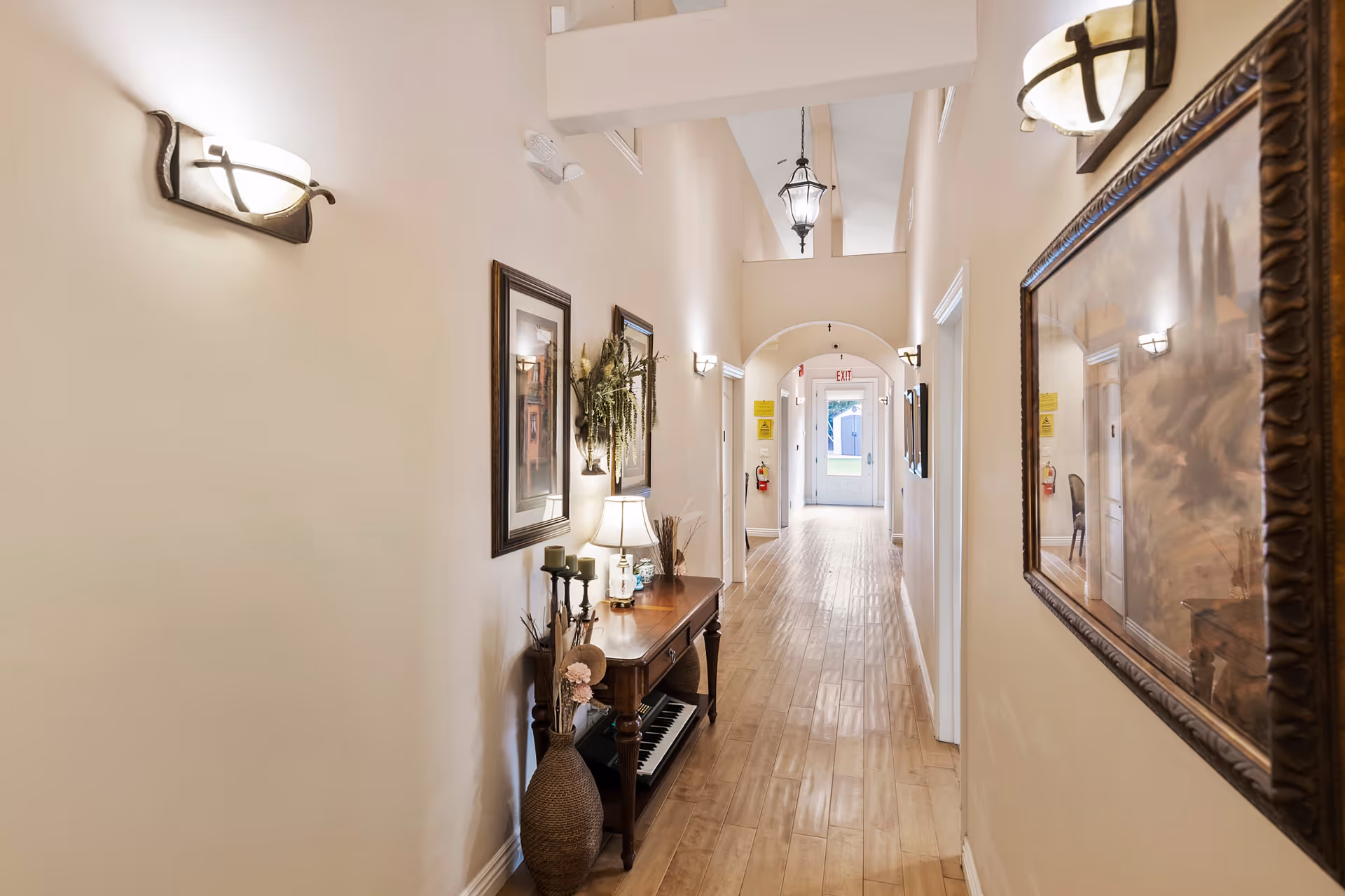 A well-lit hallway in a senior living facility with wooden flooring, beige walls, and decorative wall sconces. The hallway features framed artwork, a wooden console table with a lamp, candles, and a vase with dried flowers. At the end of the hallway, there is a door with an exit sign above it.