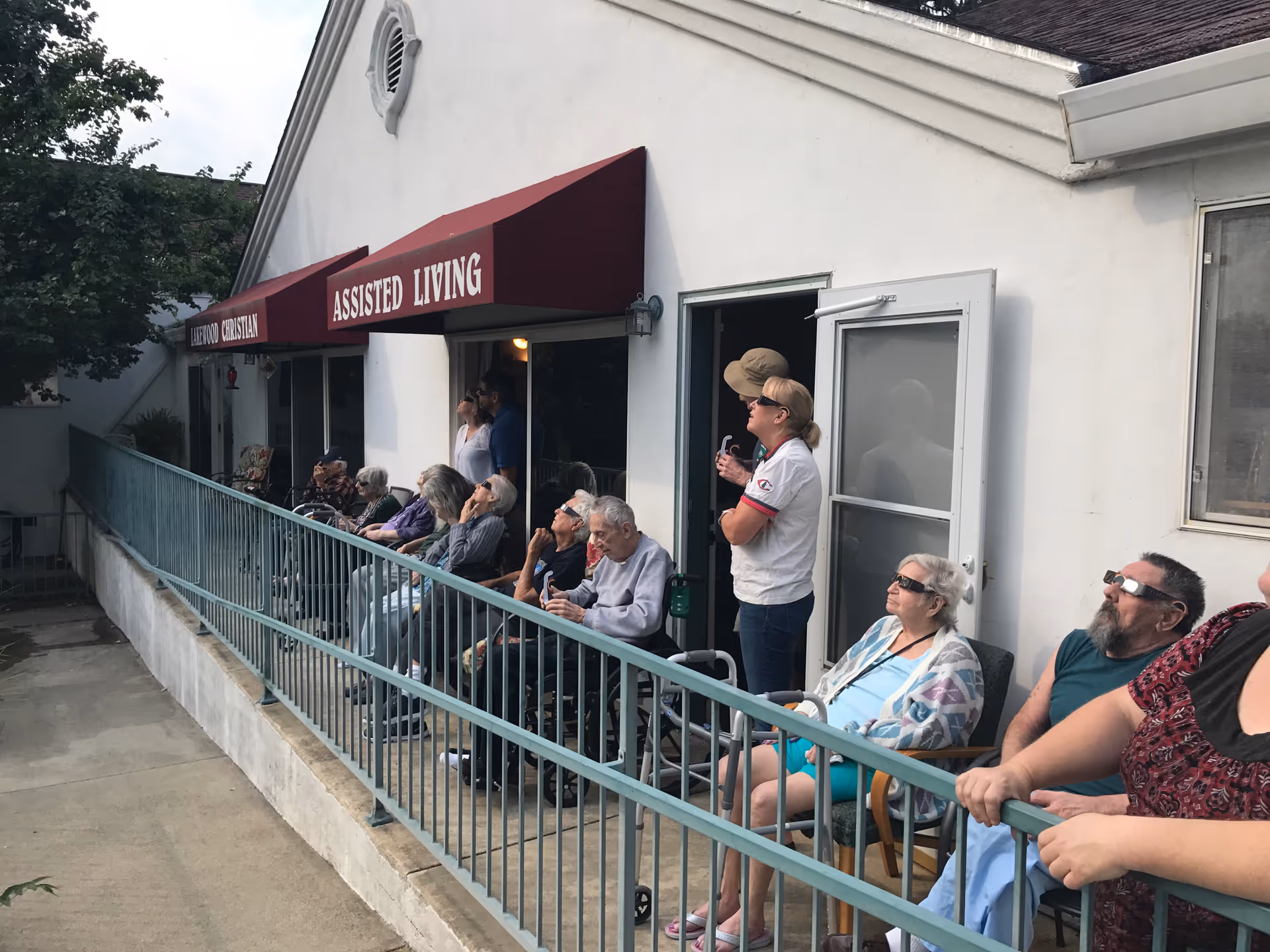 A group of elderly people and a few caregivers sitting and standing outside a building under a maroon awning that reads 'ASSISTED LIVING'. They are all wearing protective glasses and looking upwards, possibly watching an event in the sky. The building has a white exterior and a metal railing along the walkway where they are seated.