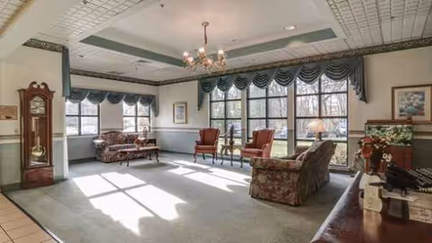 Senior living lounge with sofas, chairs, chandelier, large windows, and patterned drapes.