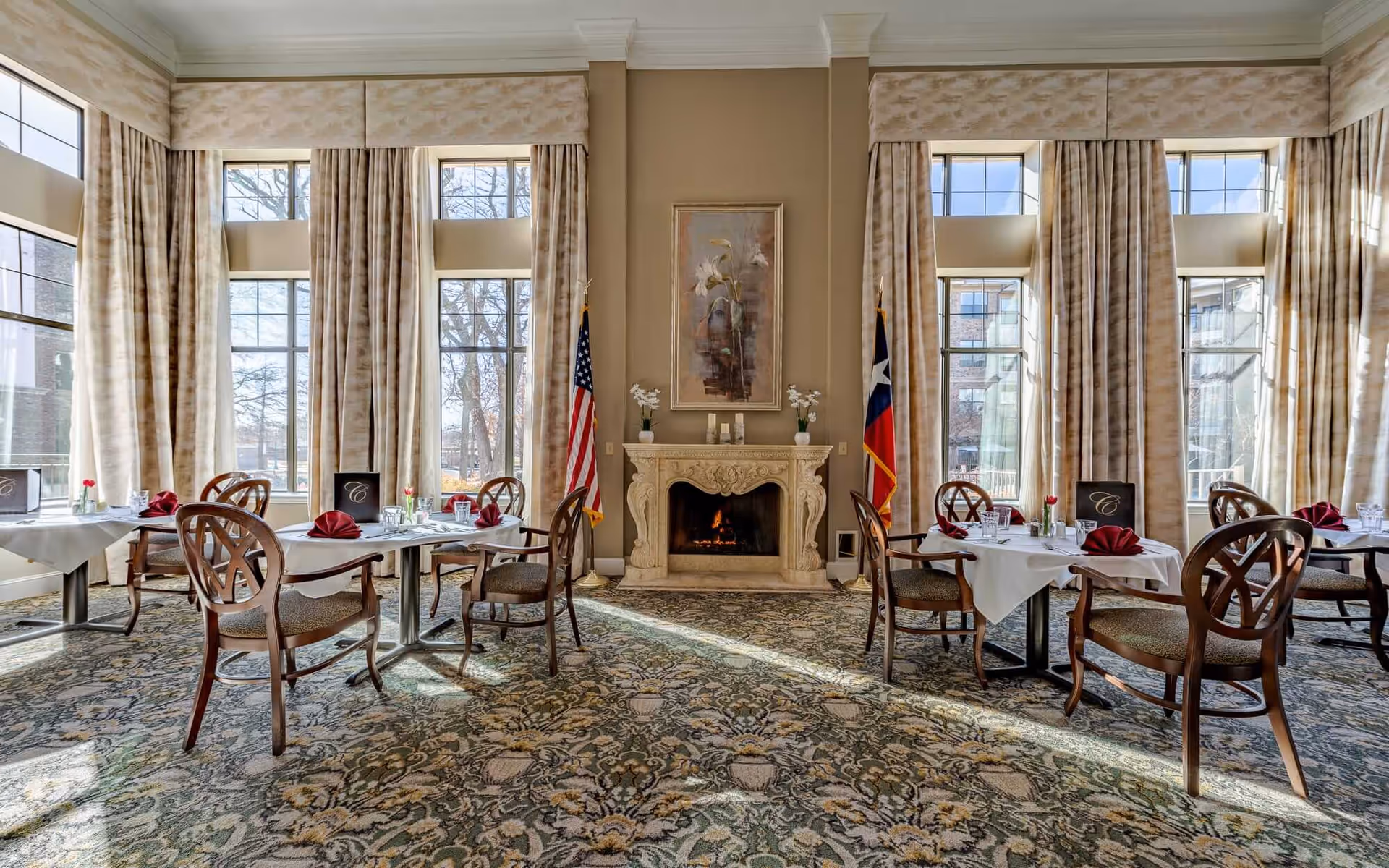 A bright dining room with large windows covered by beige curtains, allowing natural light to fill the space. The room features several tables set with white tablecloths, red folded napkins, glassware, and menus. There are wooden chairs with cushioned seats around the tables. In the center of the back wall is a decorative fireplace with a floral painting above it, flanked by an American flag on the left and a Texas state flag on the right. The floor is covered with a patterned carpet.