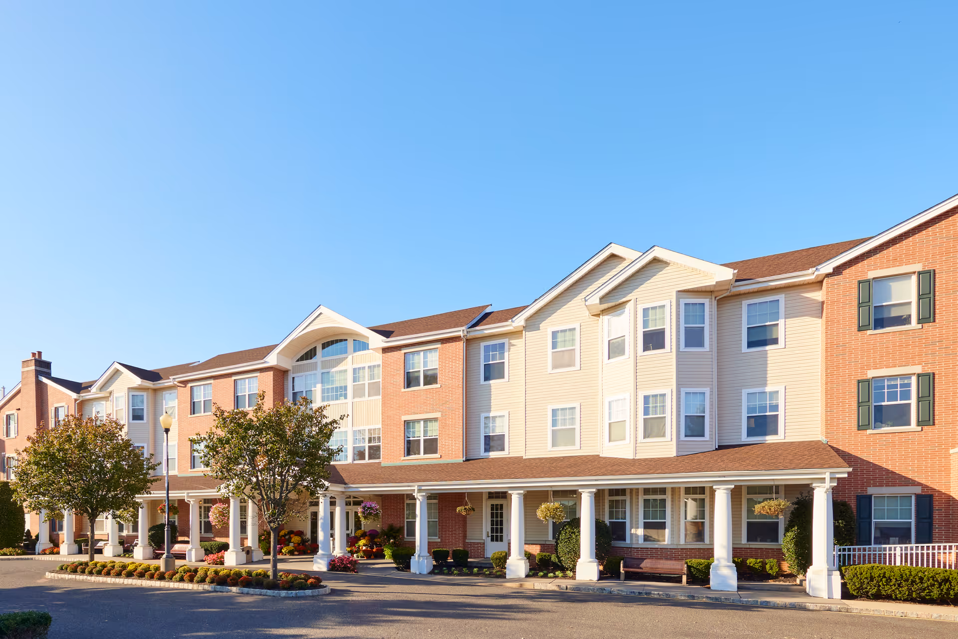 Front exterior of a three-story brick-and-siding senior living building with a covered porch, columns, and landscaped entrance under a clear blue sky.