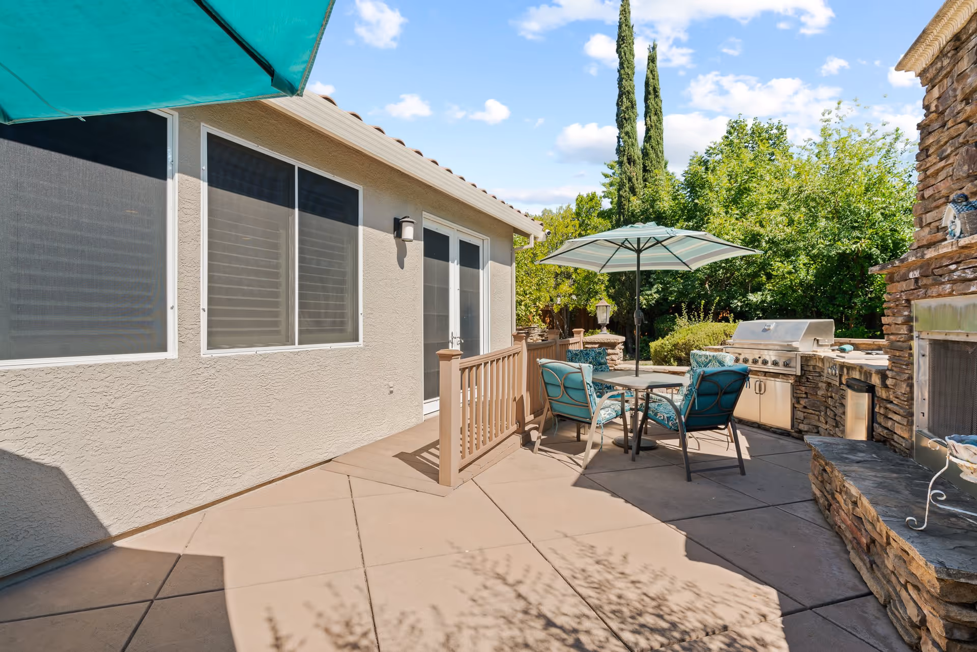Outdoor patio area with a round table and four cushioned chairs under a large umbrella. There is a built-in stone grill and countertop on the right side, surrounded by greenery and trees under a blue sky with scattered clouds.