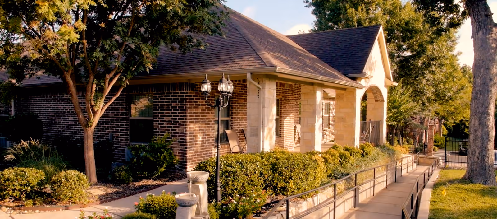 Exterior view of a brick and stone building with a sloped roof, surrounded by well-maintained bushes, trees, and a paved walkway with a handrail leading to the entrance. There is a vintage-style street lamp and a small water fountain in the garden area.