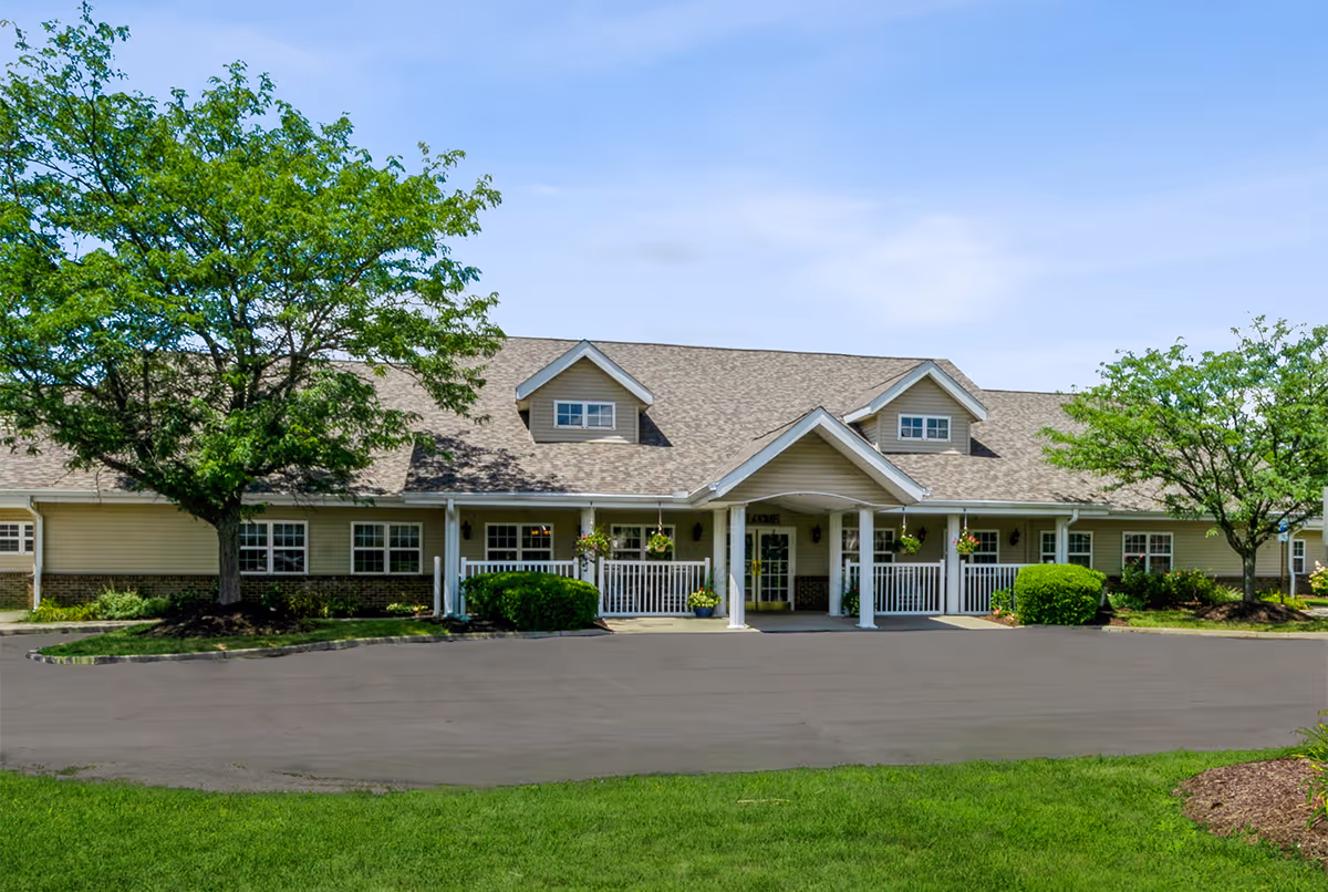 Front exterior view of a single-story building with beige siding and a shingled roof, featuring a covered entrance supported by white columns. The building is surrounded by green grass, trees, and shrubs under a clear blue sky.